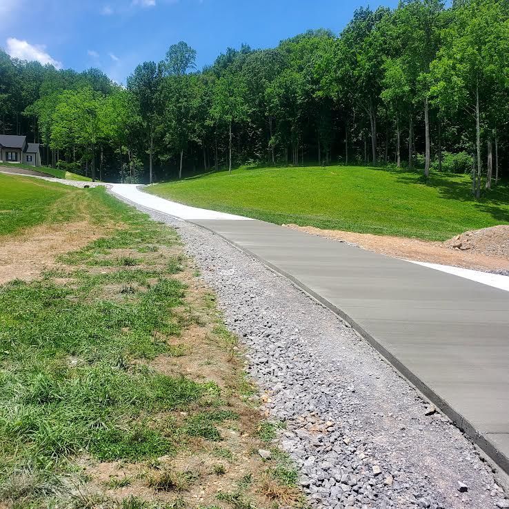 Newly poured concrete driveway curves up a grassy hill towards a house and trees under a blue sky.