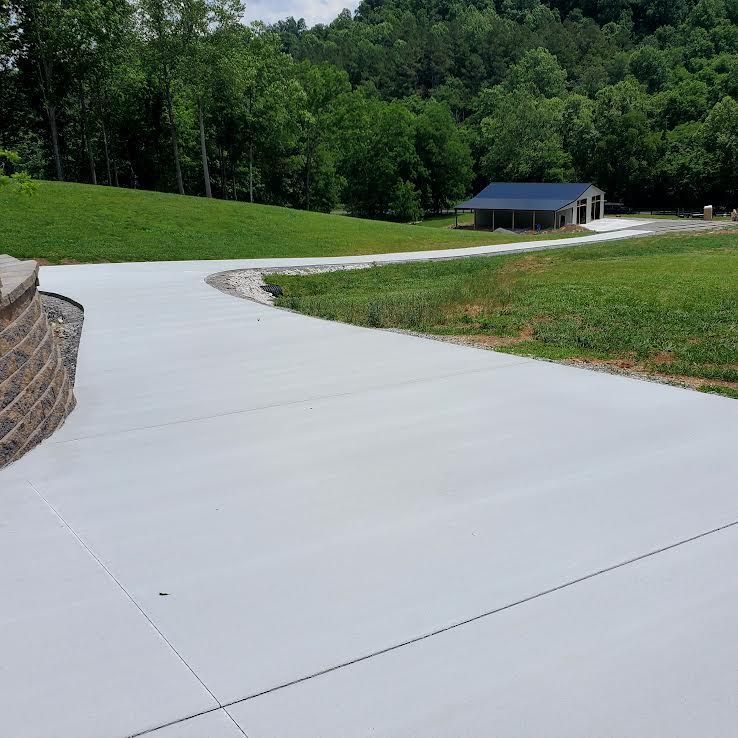 Concrete driveway curves toward a building with a blue roof, surrounded by green grass and trees.