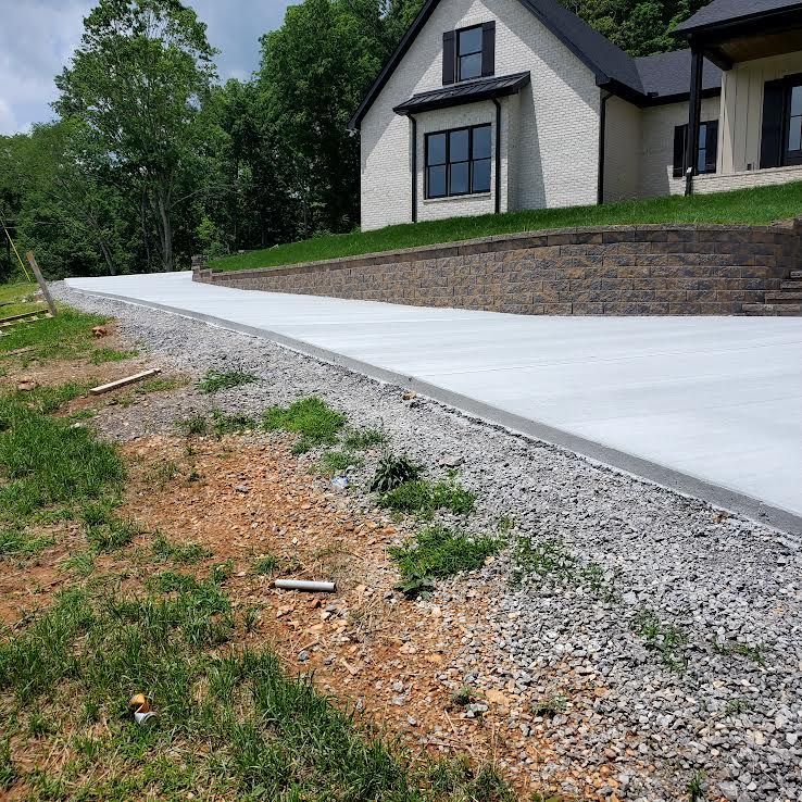 A newly poured concrete driveway leads uphill to a white brick house with black trim, built on a graded lot.