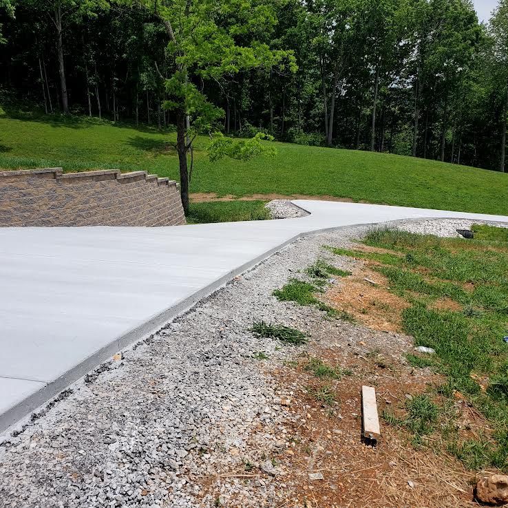 A newly poured concrete driveway curves toward a grassy hill and trees, with gravel edges.
