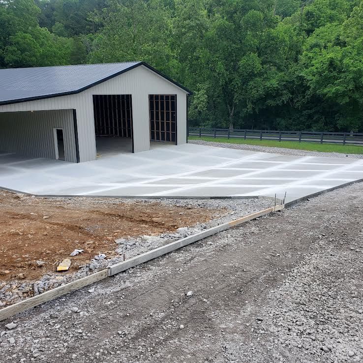 Newly poured concrete driveway with a metal-roofed garage in the background. Gravel and dirt border the concrete.