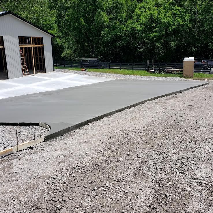 Newly poured concrete slab in front of a barn, on a gravel surface. Construction in progress outdoors.