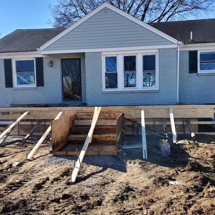 House under construction: wooden steps lead to a raised porch. Gray house with black shutters, blue siding.