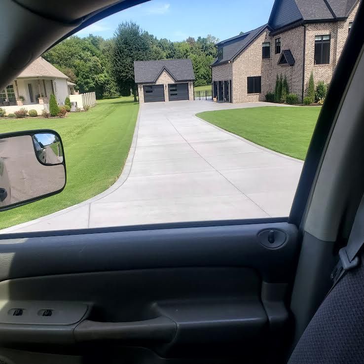 View from a car window of a long concrete driveway leading to a large house with a detached garage on a sunny day.