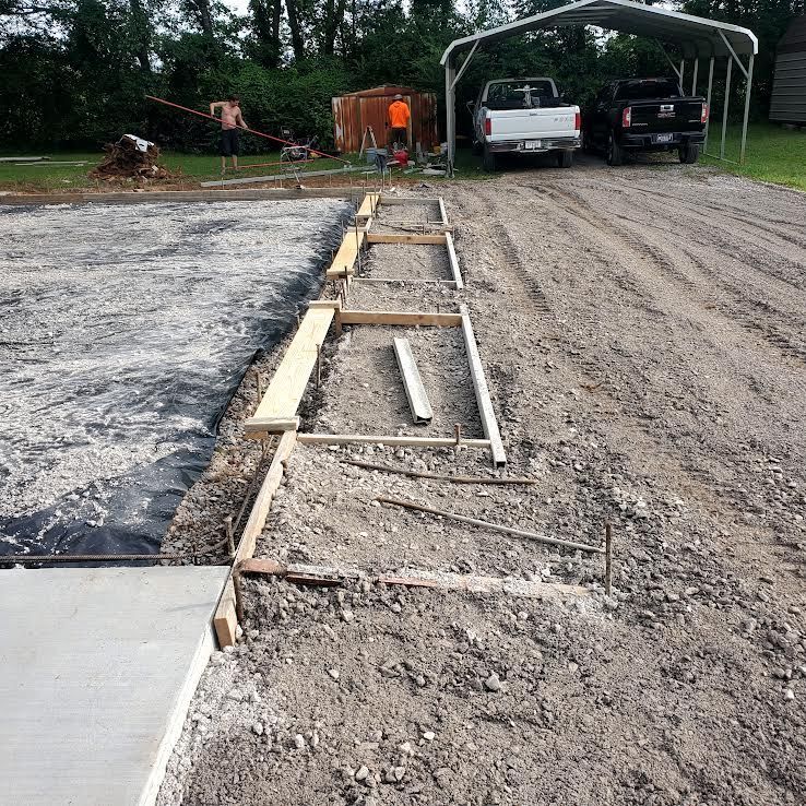 Construction site: Wooden forms for concrete work; vehicles parked nearby; worker in orange shirt in the distance.