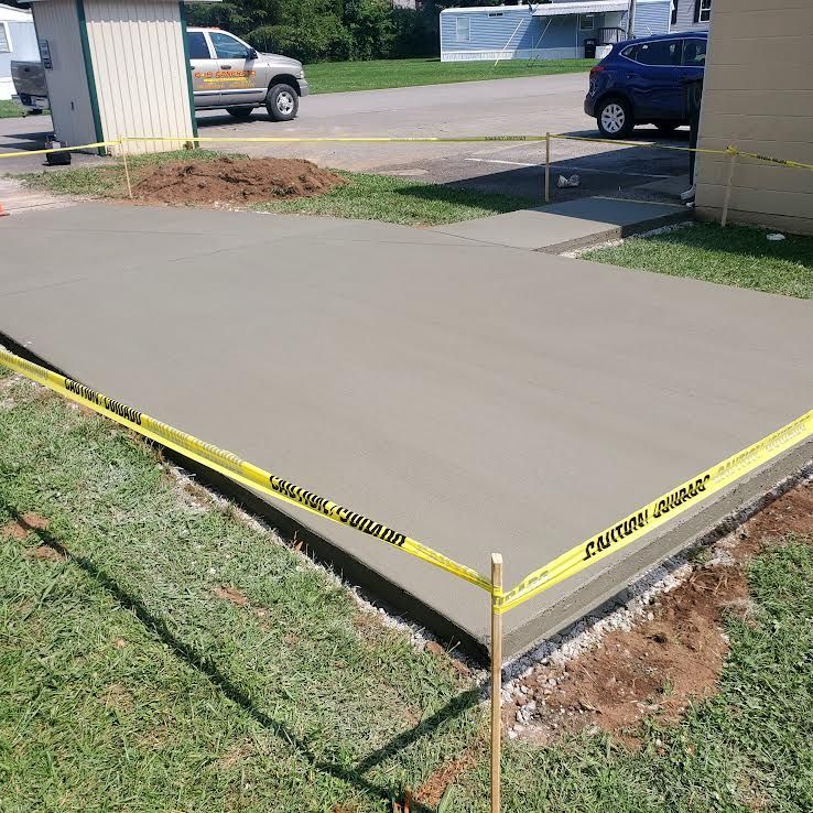 Freshly poured concrete slab bordered by yellow caution tape. Grass and a sidewalk are visible.