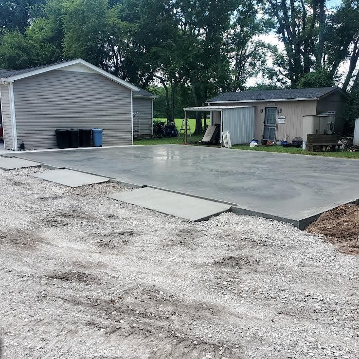 Newly poured concrete driveway with gravel bordering, next to a garage and sheds.