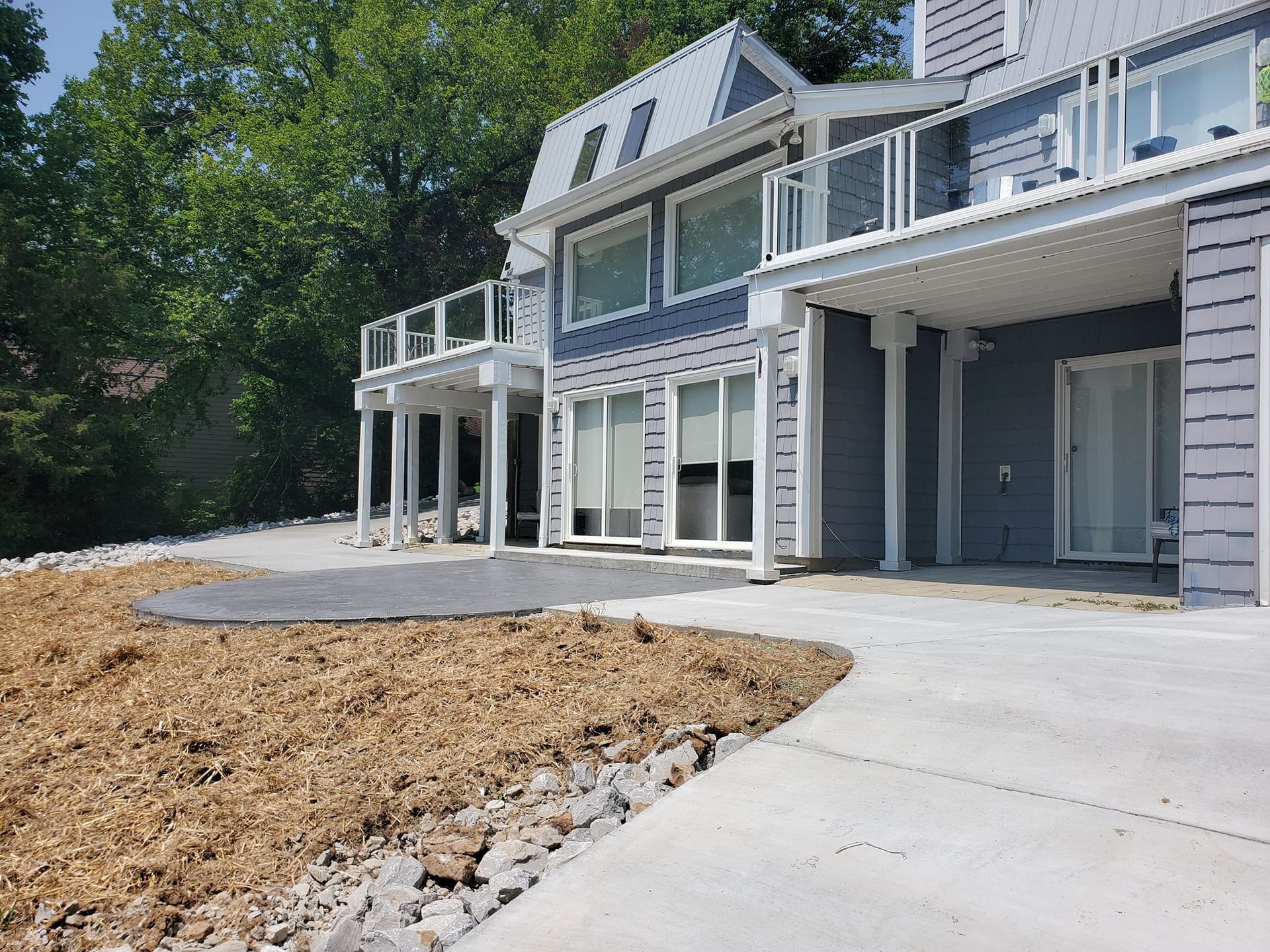 House with blue siding, two-story deck, and concrete patio. Yard under construction with wood chips and rocks.