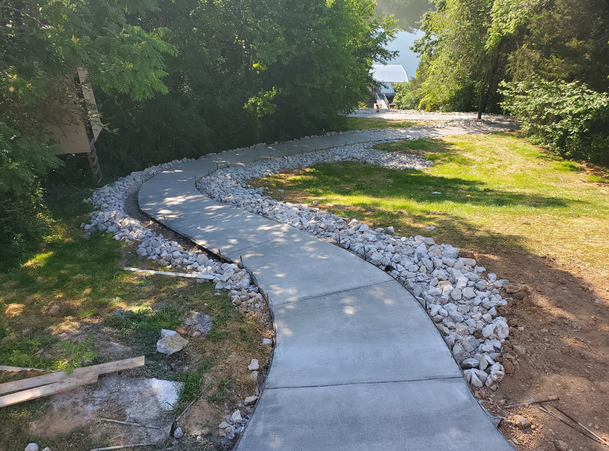 Curving paved pathway bordered by rocks, leading through grassy area with trees.