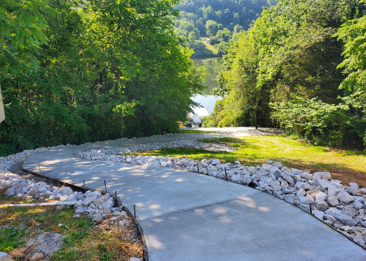Concrete pathway leading downhill, lined with rocks, to a lake surrounded by trees.