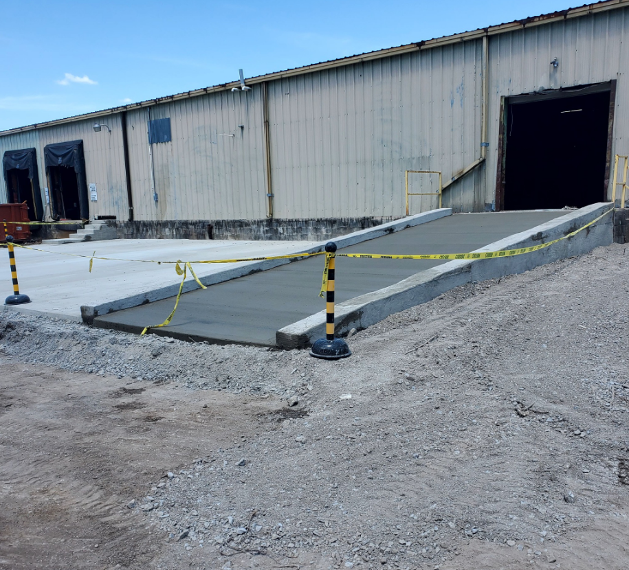 Concrete ramp leading to a warehouse loading dock, bordered by concrete walls, caution tape, and safety posts.