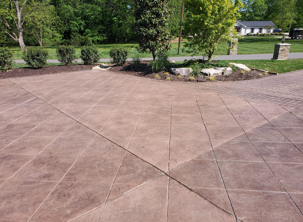 Brown patterned concrete patio with planted borders, trees, and a house in the background.