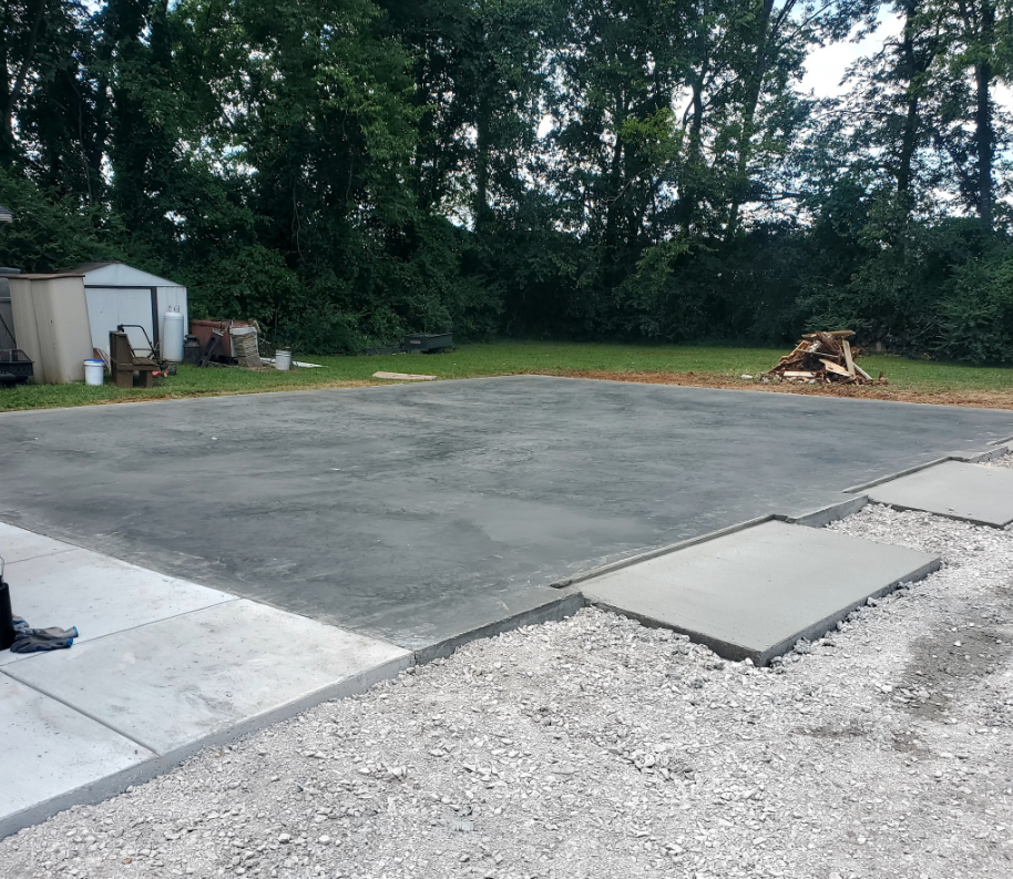 Concrete pad with gravel border in a grassy yard, with trees in the background.