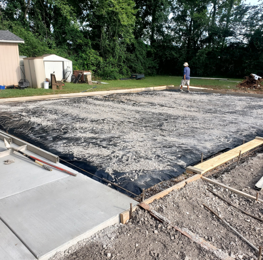 Construction site with concrete forms. A worker stands in the background. Gravel and black sheeting cover the ground.