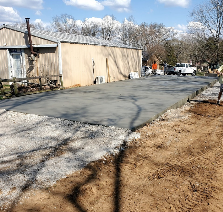Newly poured concrete slab next to a building. Gravel driveway and dirt foreground.
