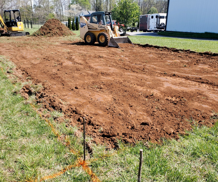 A skid steer leveling dirt in a rectangular area on a grassy lawn; a small excavator and dirt pile are visible.