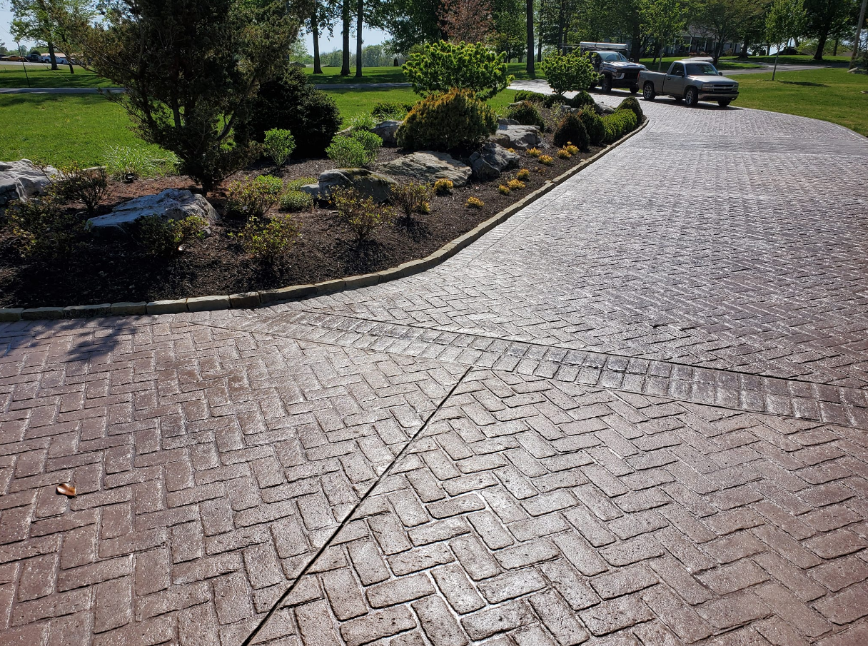 Brick-patterned driveway curves past a landscaped garden with vehicles parked in the background.