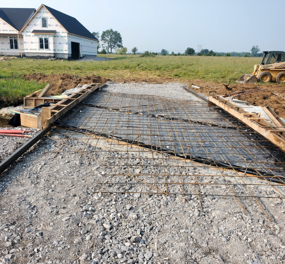 Construction site: gravel base with rebar grid laid for a concrete driveway. A house is in the background.