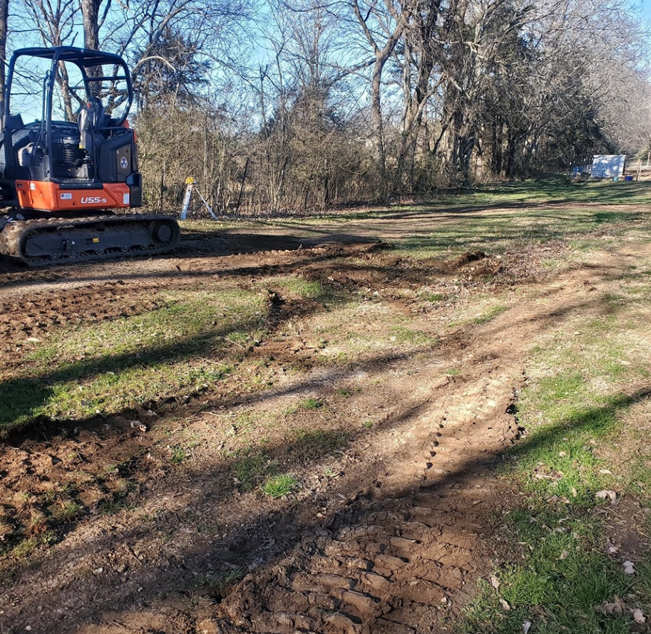 Mini excavator on a grassy area, creating tracks. Trees in the background, sunny day.
