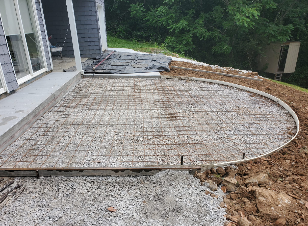 Concrete patio construction with gravel base and rebar grid, next to a house with a curved edge.
