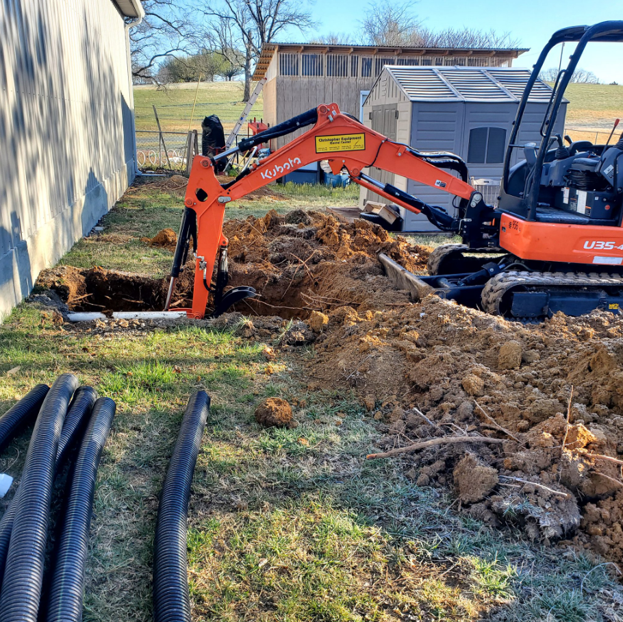 Orange Kubota excavator digging a trench near a building. Black pipes lie on the grass.