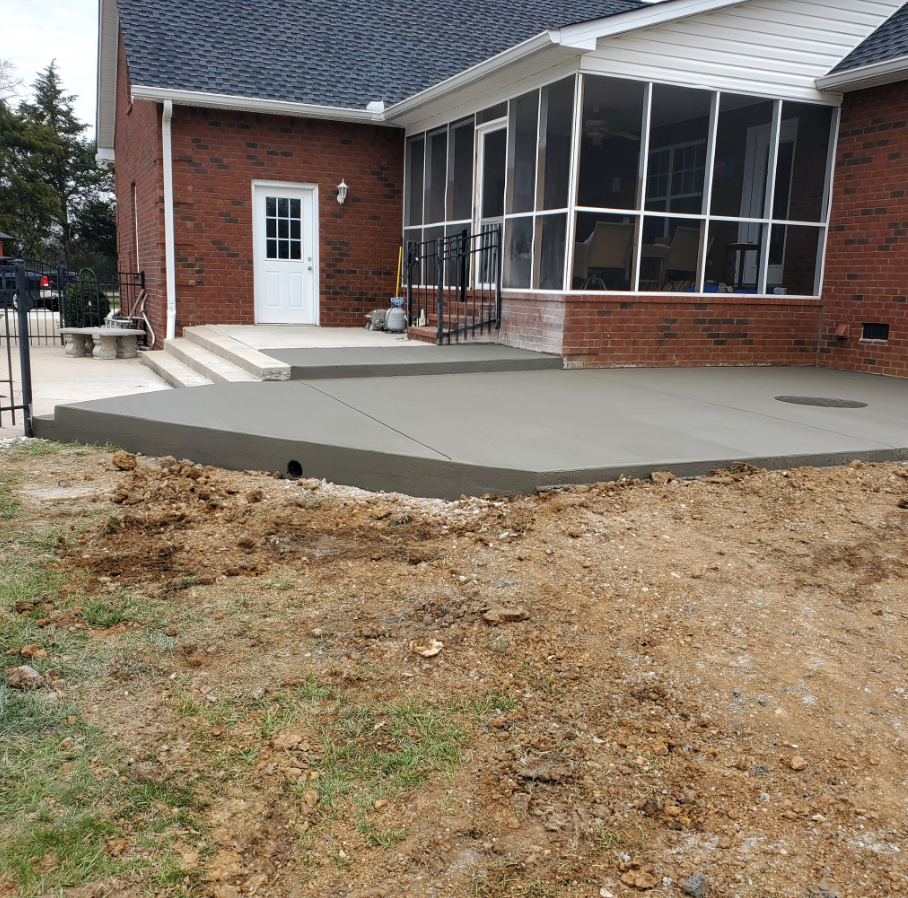 Newly poured concrete patio beside a brick house with a screened porch and white door.