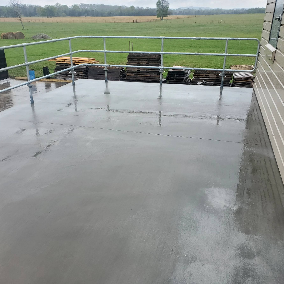 A newly poured concrete patio with a metal railing. A grassy field and cloudy sky are in the background.