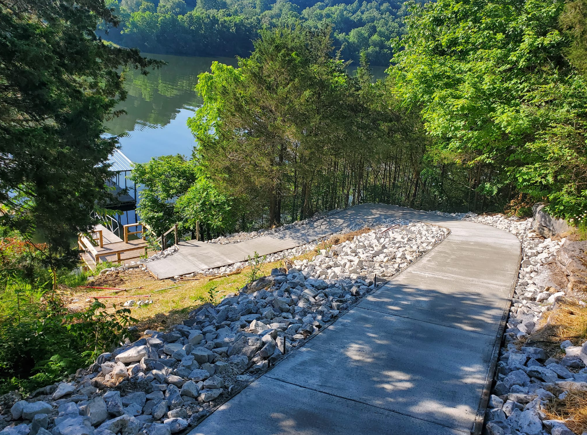 Concrete walkway down a hillside, bordered by rocks, leading to a body of water with trees.