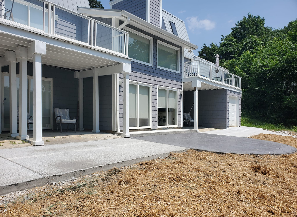 Blue beach house with multiple levels, porch, and a stone patio. Overgrown with woodchips. Sunny day.