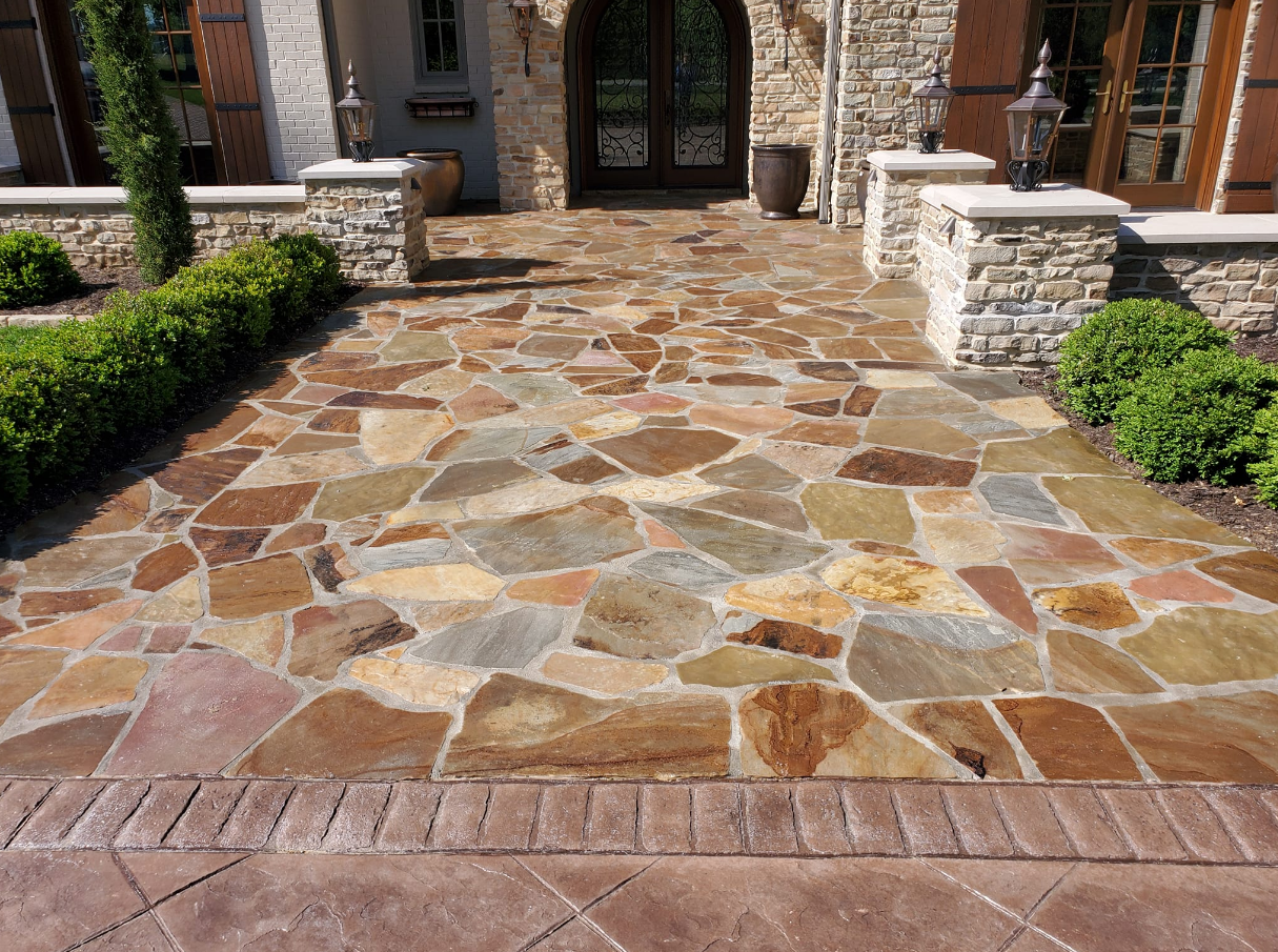 Stone path leading to a home's arched front door. Path is bordered by brick and stone columns with greenery.