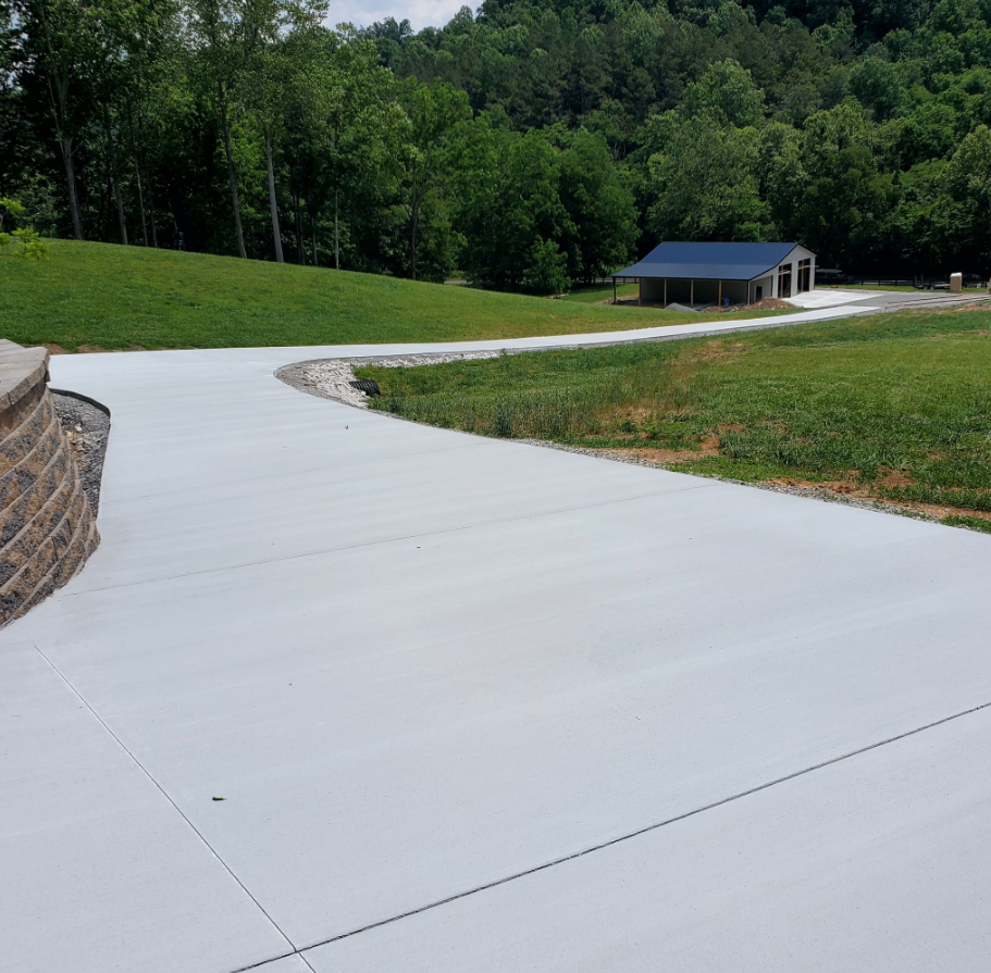Concrete driveway curves toward a building with a blue roof, surrounded by green grass and trees.