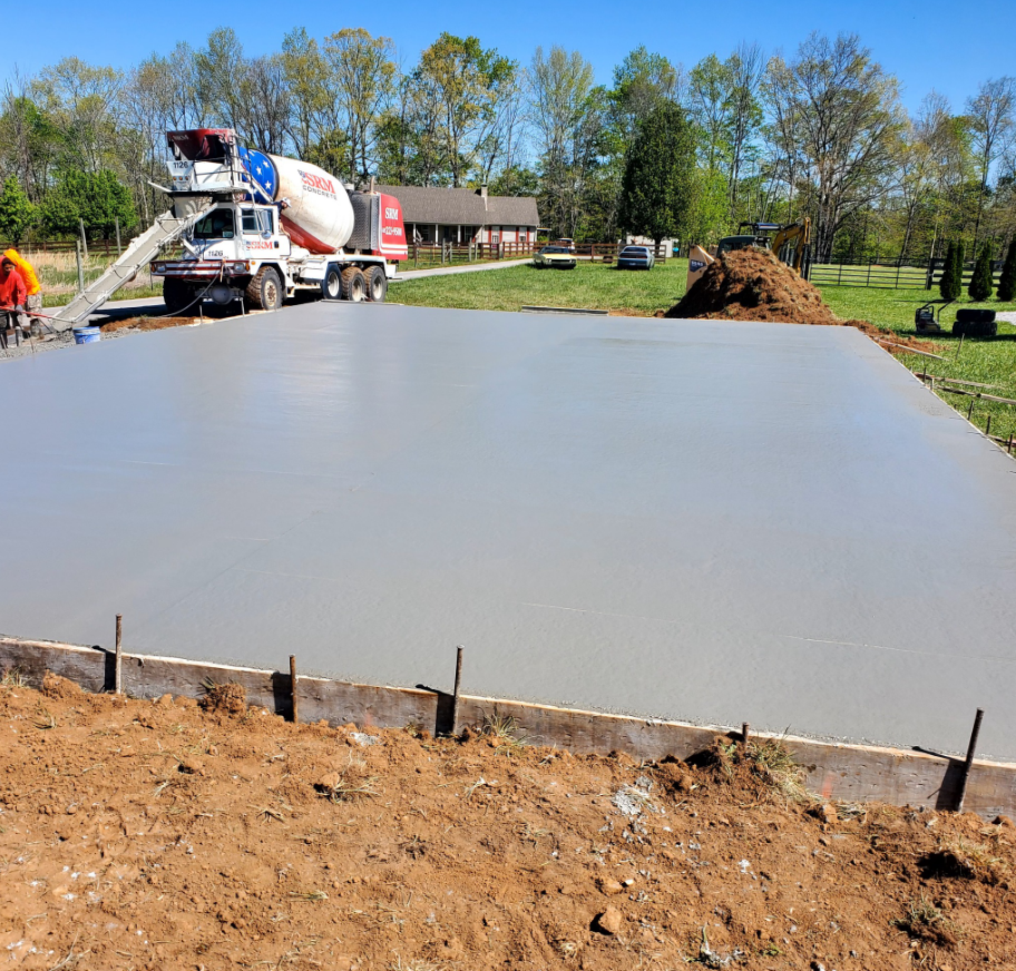 Concrete being poured from a truck, forming a smooth gray slab within wooden forms on a dirt lot.