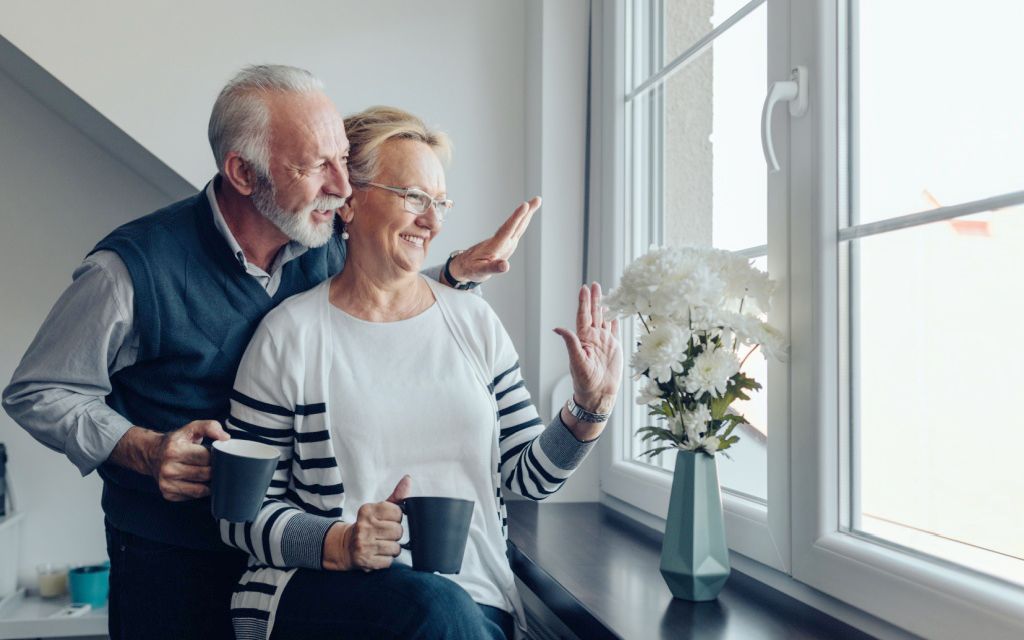 An elderly couple, smiling, looks out a window, waving, with mugs. The man's arm rests on the woman's shoulder.