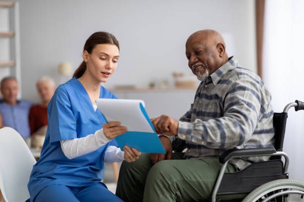 A healthcare worker in blue scrubs reviews paperwork with an elderly Black man in a wheelchair, likely in a care facility. Other residents are visible in the background.