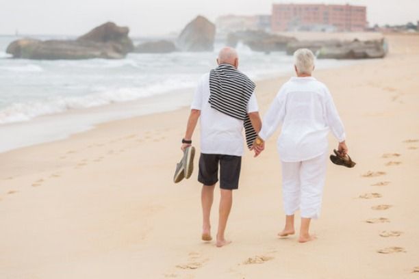 Elderly couple holding hands, walking barefoot on a sandy beach towards the ocean, carrying sandals, with a large rock formation in the background.