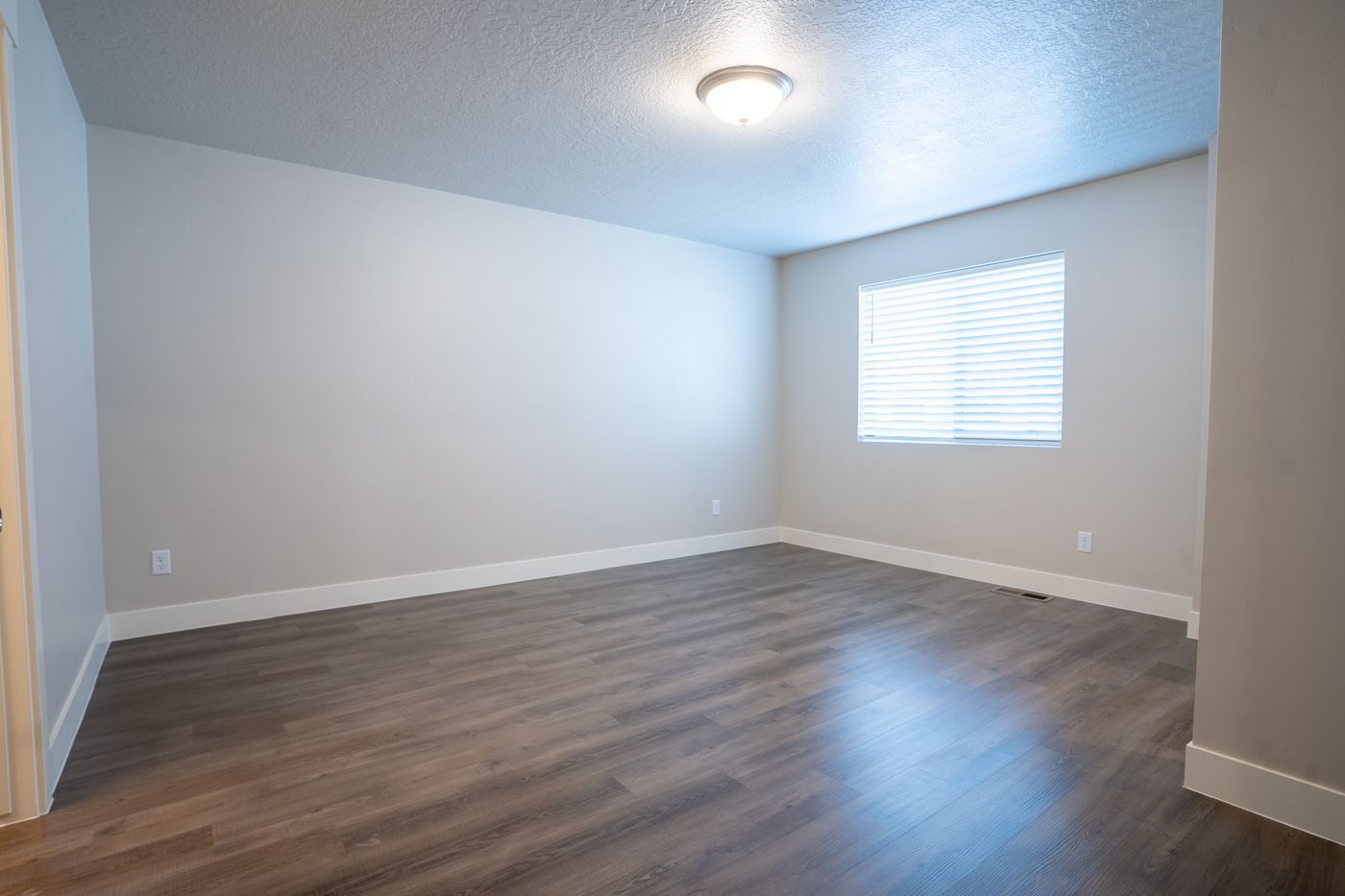 Empty living room with gray walls, dark wood floors, baseboards, and a window with blinds.