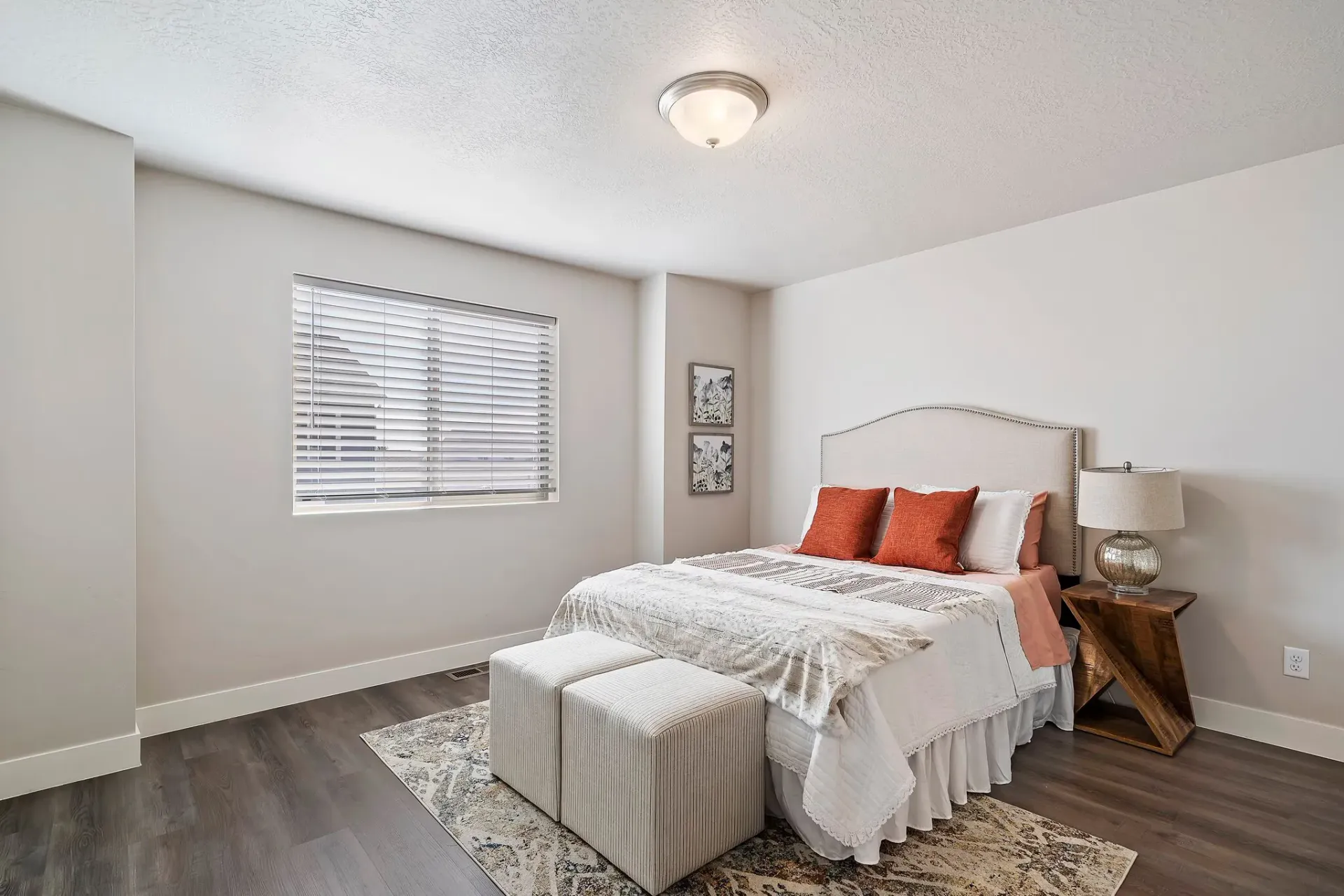 Bedroom in an apartment with a beige upholstered headboard, two ottoman benches, nightstand, lamp, and window with blinds.