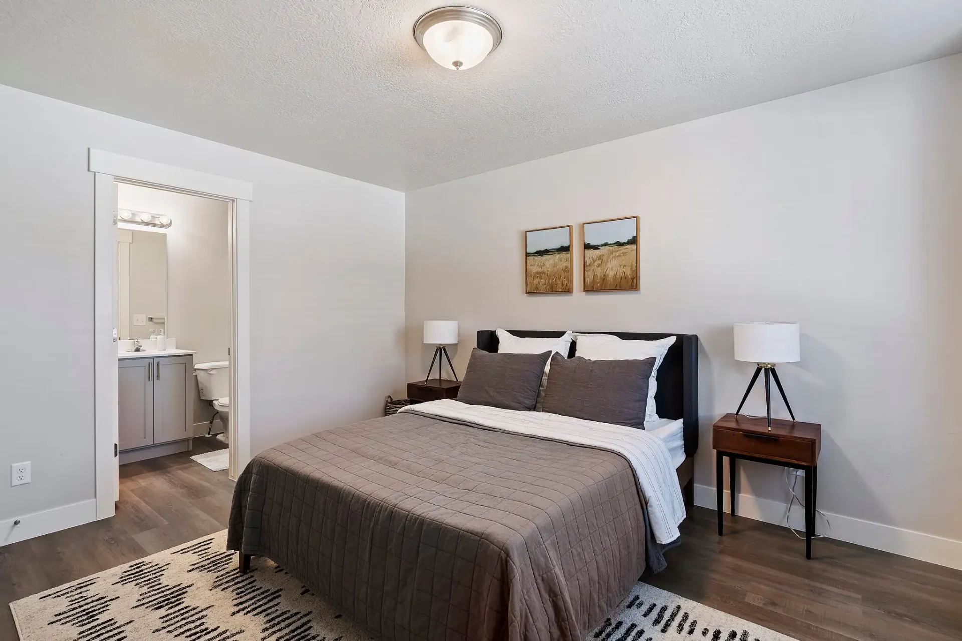 Bedroom in an apartment with a bed, two nightstands, lamps, and an open doorway to a bathroom.