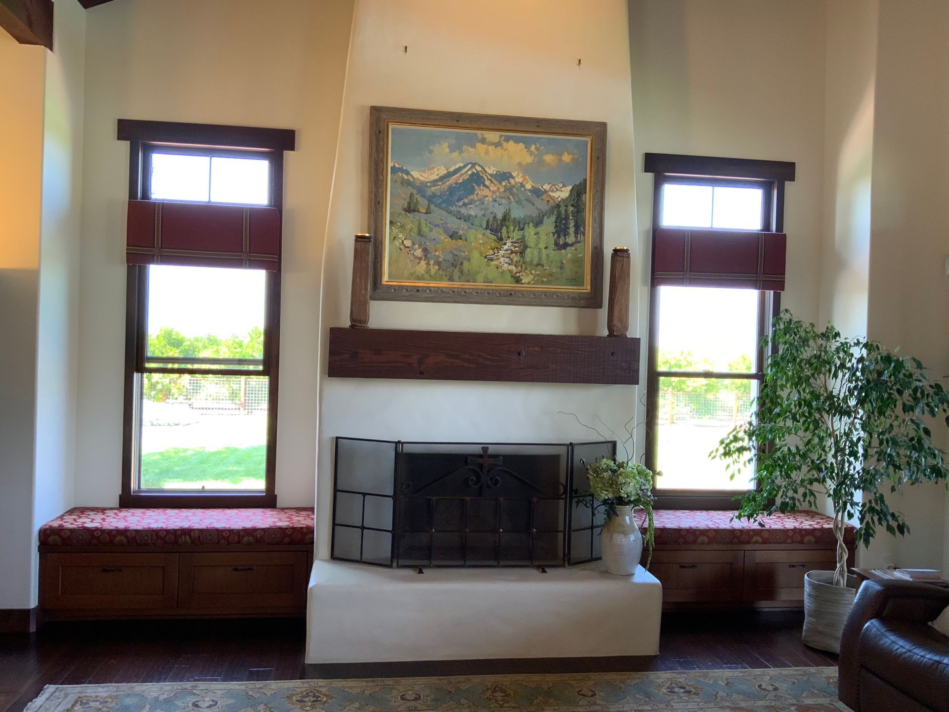 Fireplace with windows on either side, painting above. Two window seats with red accents. Green plant on the right.