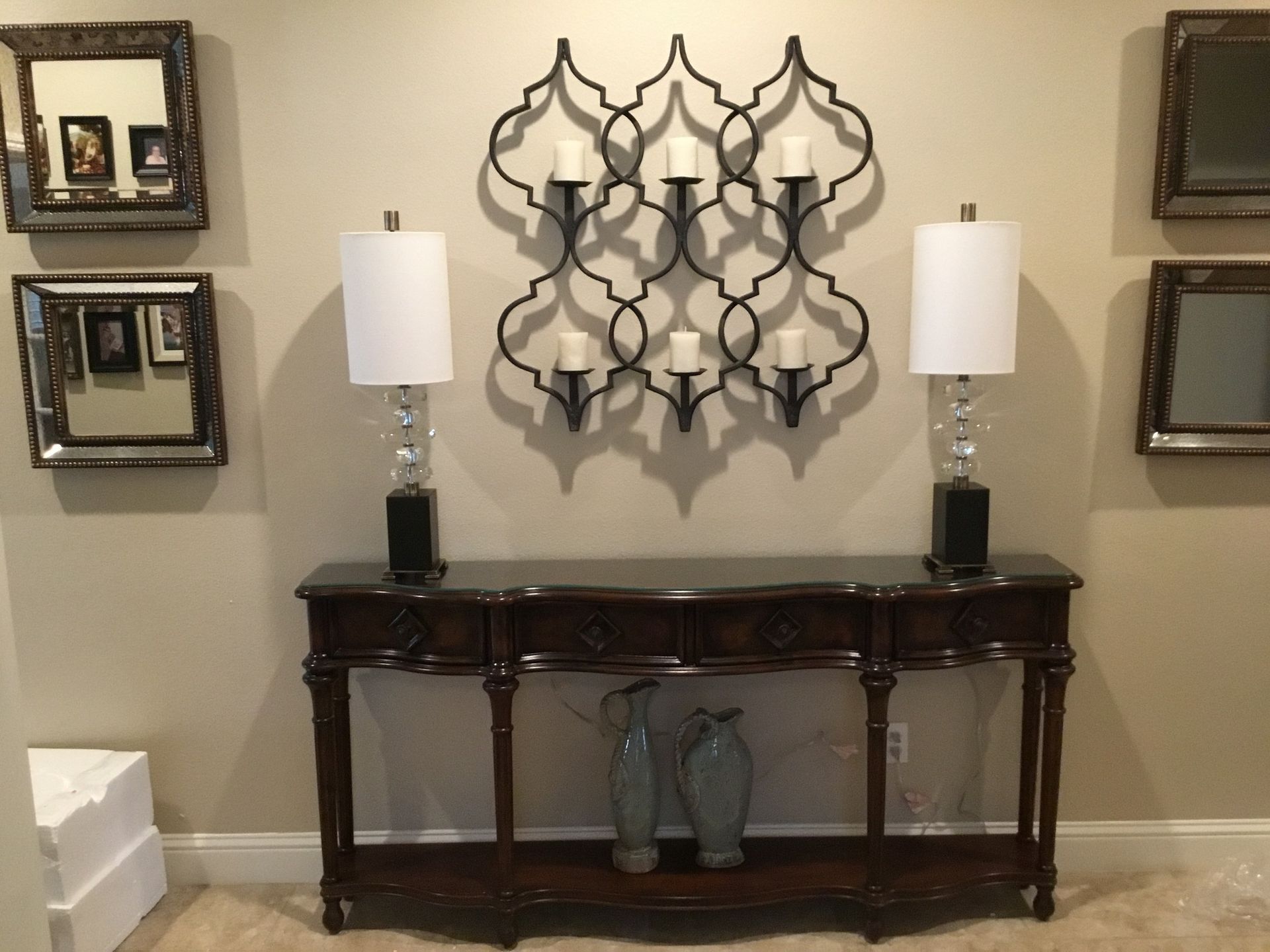 A console table with lamps, decor, and a wall of mirrors and a candle holder against a beige wall.