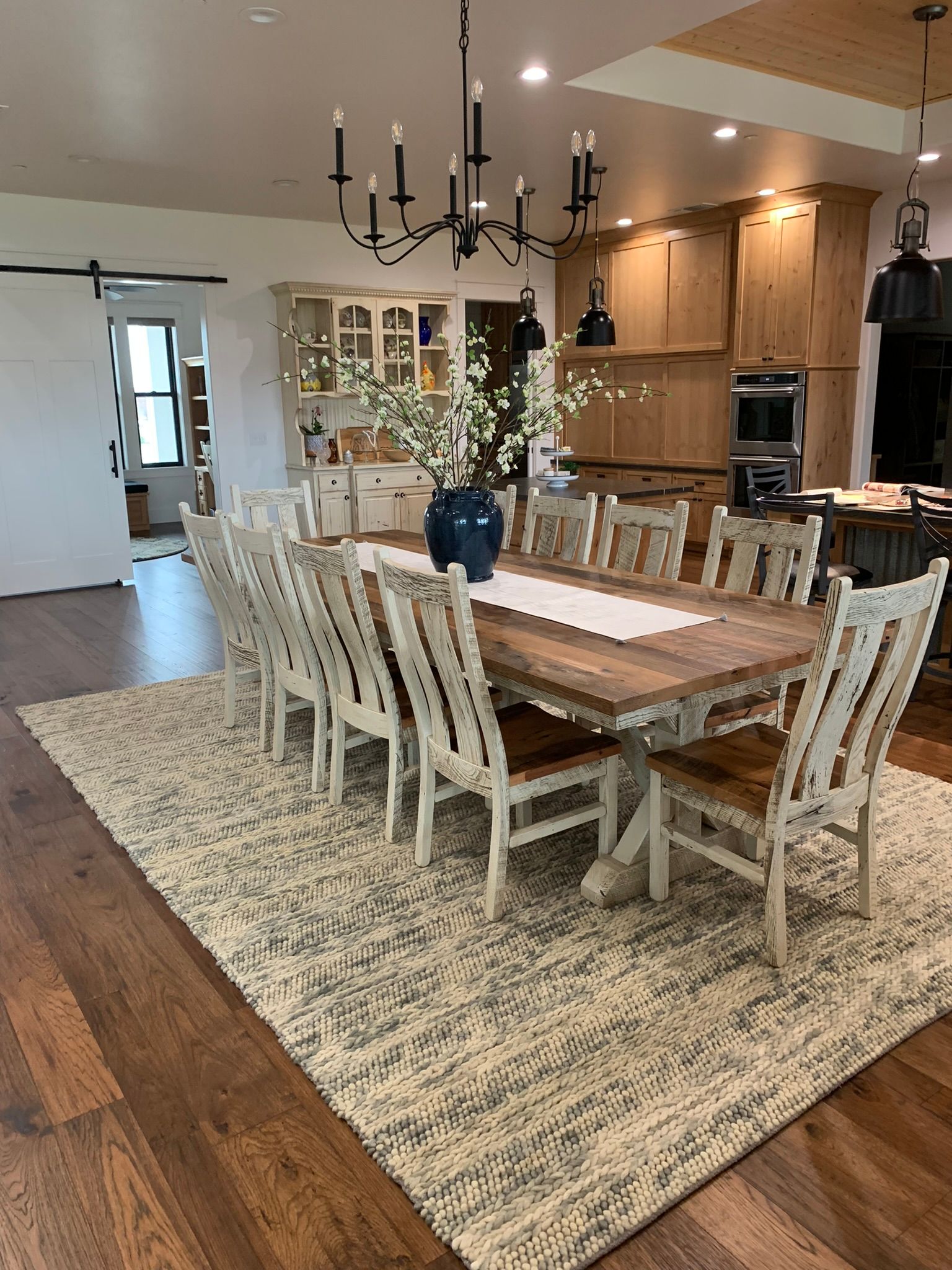 Dining room with large wooden table, chairs, rug, and chandelier. Kitchen cabinets in background.