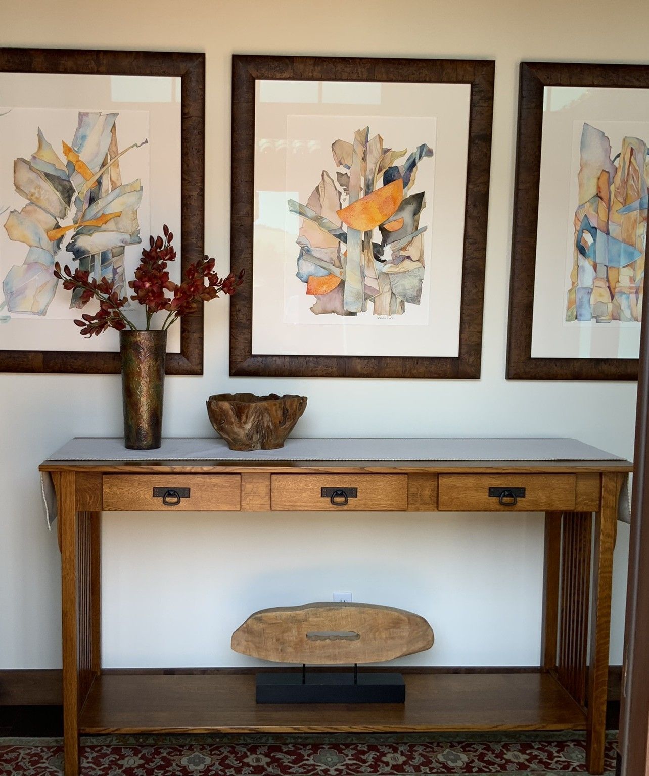 A wooden console table under three framed abstract watercolor prints, decorated with a vase of branches and a wooden bowl.