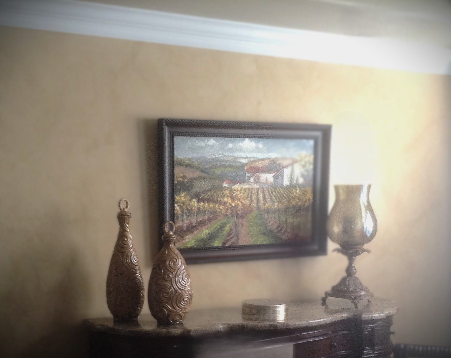 Two decorative vases and a glass lamp sit on a marble surface below a framed landscape painting on a tan textured wall.