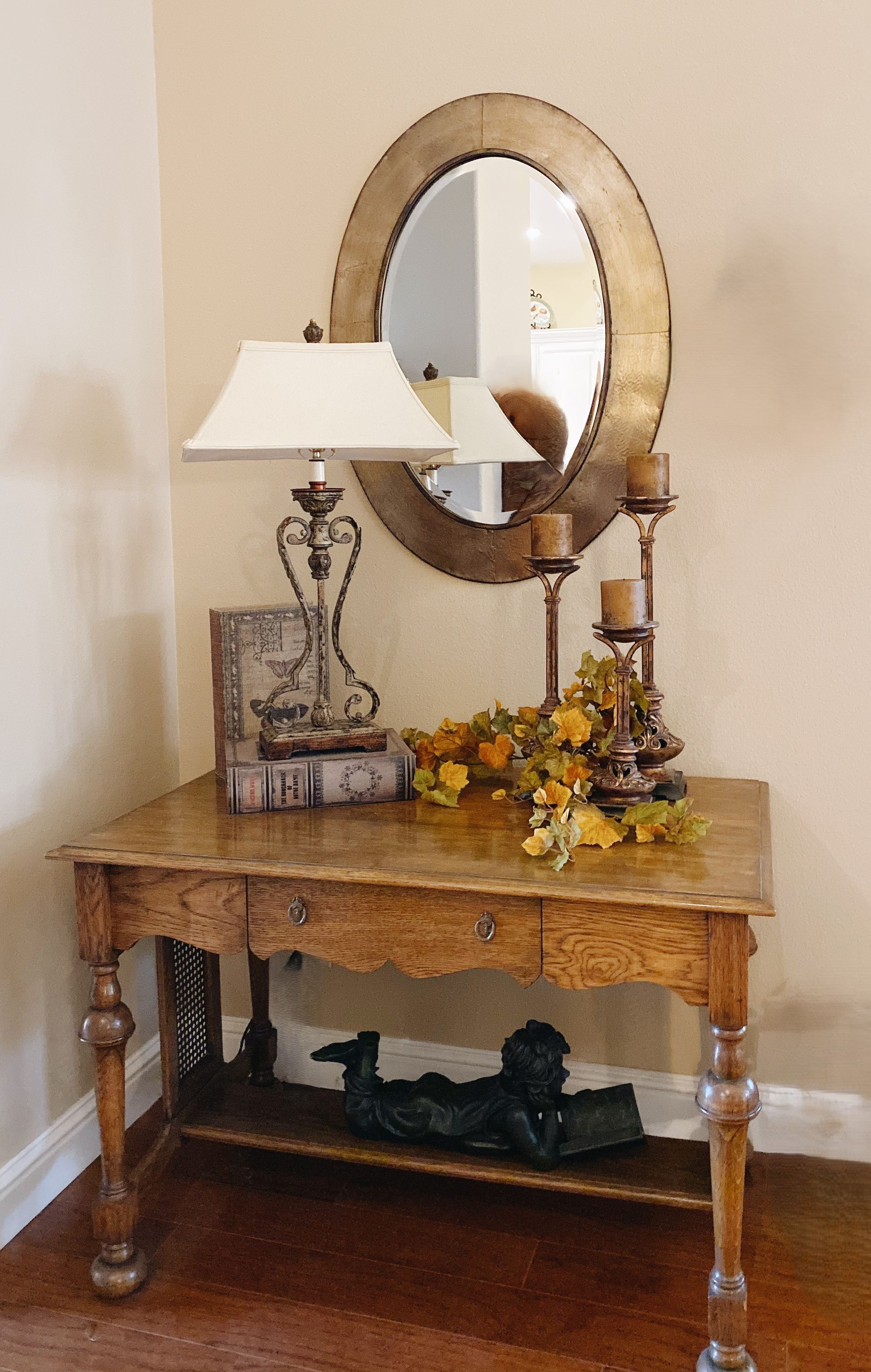 An oval mirror hangs above a wooden console table with a lamp, candles, fall foliage, and a figurine on the lower shelf.