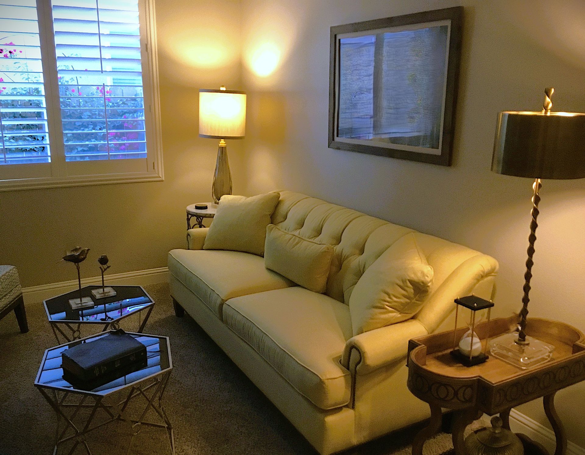 A tufted yellow sofa in a living room with two end tables, two lamps, a mirror, and stacked hexagonal coffee tables.