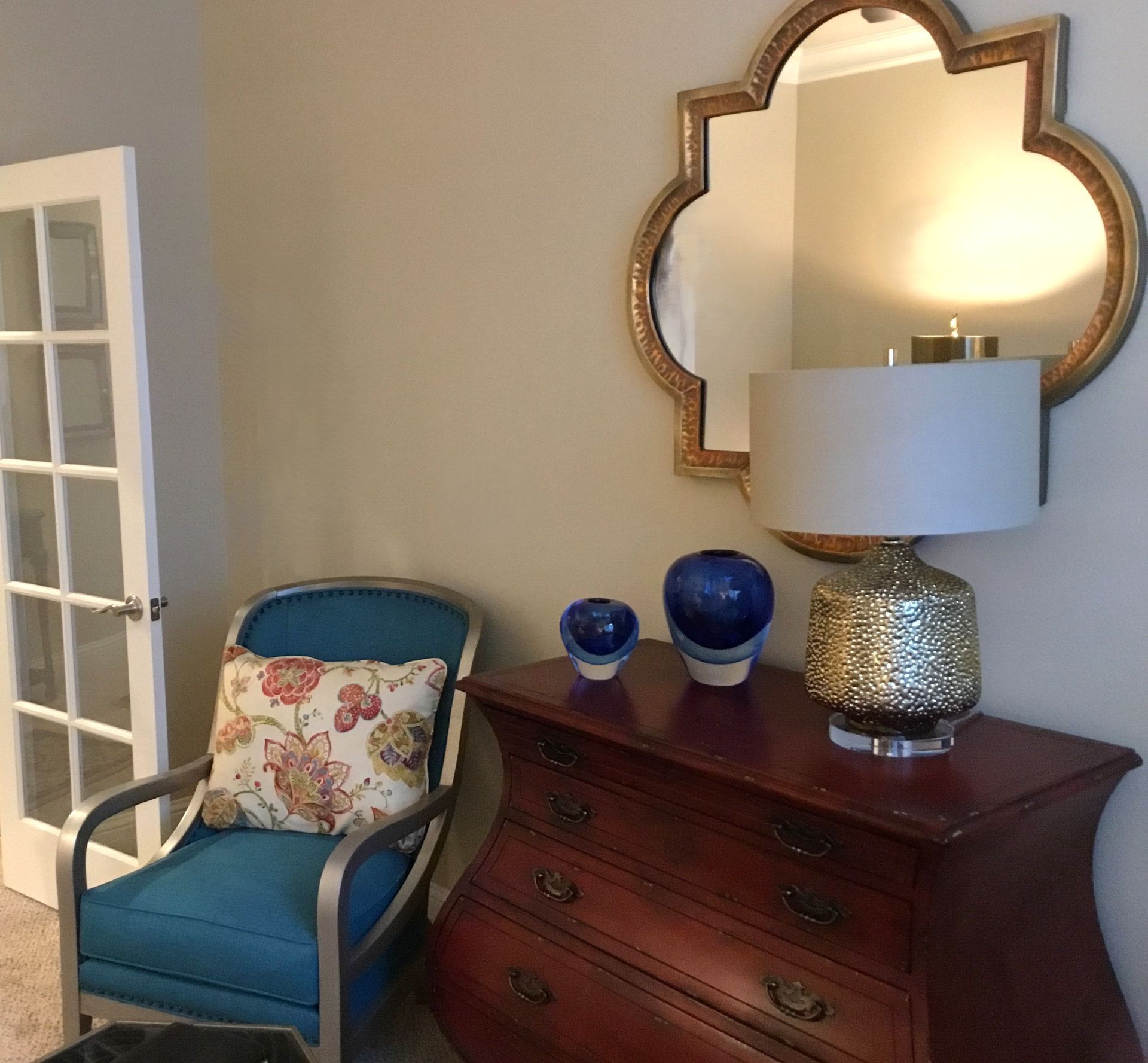 A teal armchair with a floral pillow sits next to a dark wood dresser with two blue vases, a lamp, and a wall mirror.