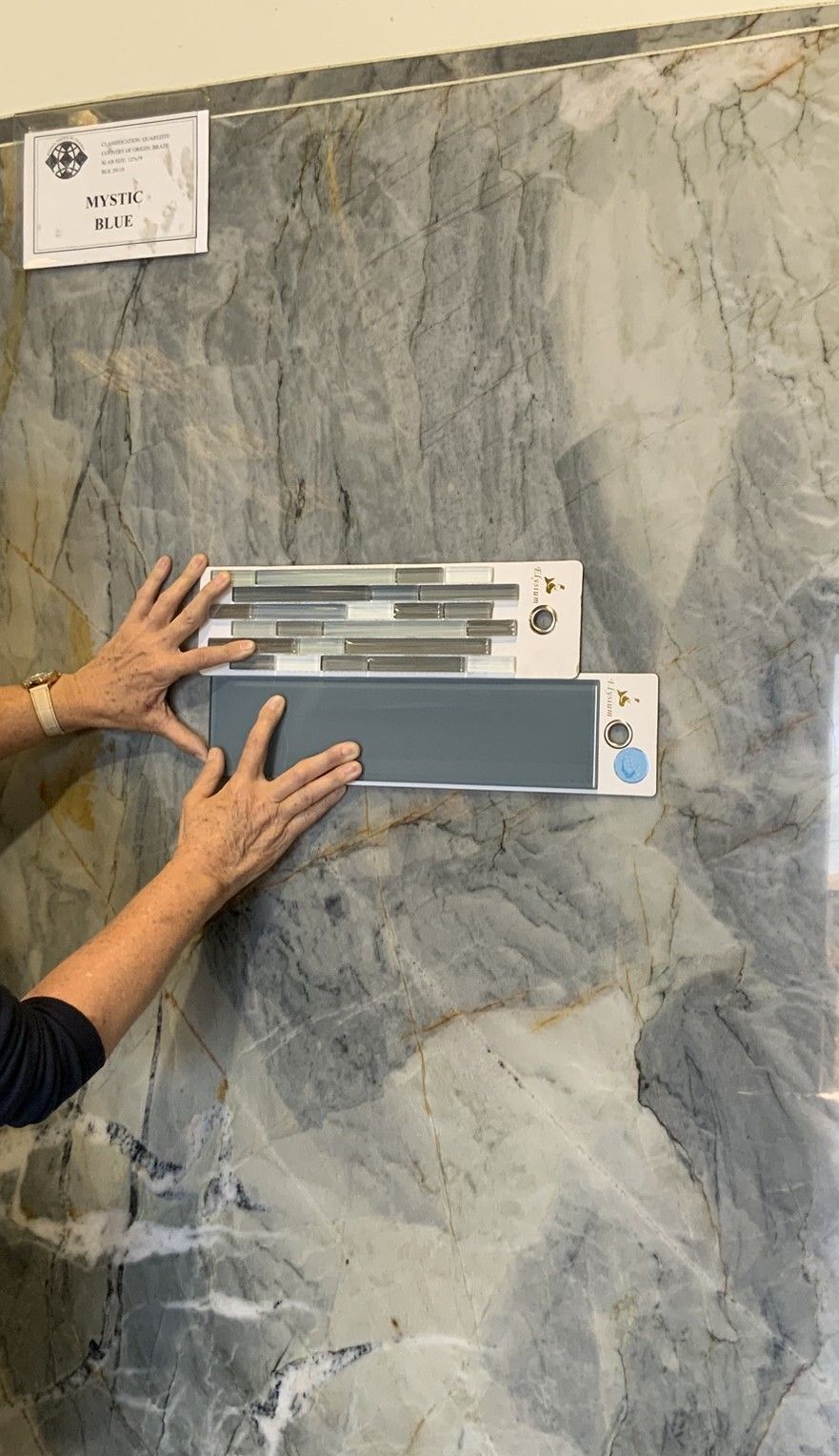 Hands hold two tile samples against a large, grey, veined marble slab in a showroom.