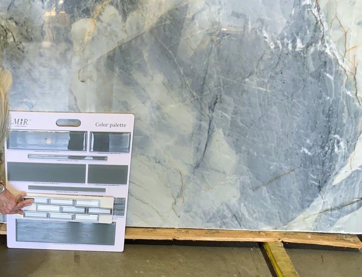 A hand holds a sample board of grey tiles against a large, polished slab of white and grey marble.