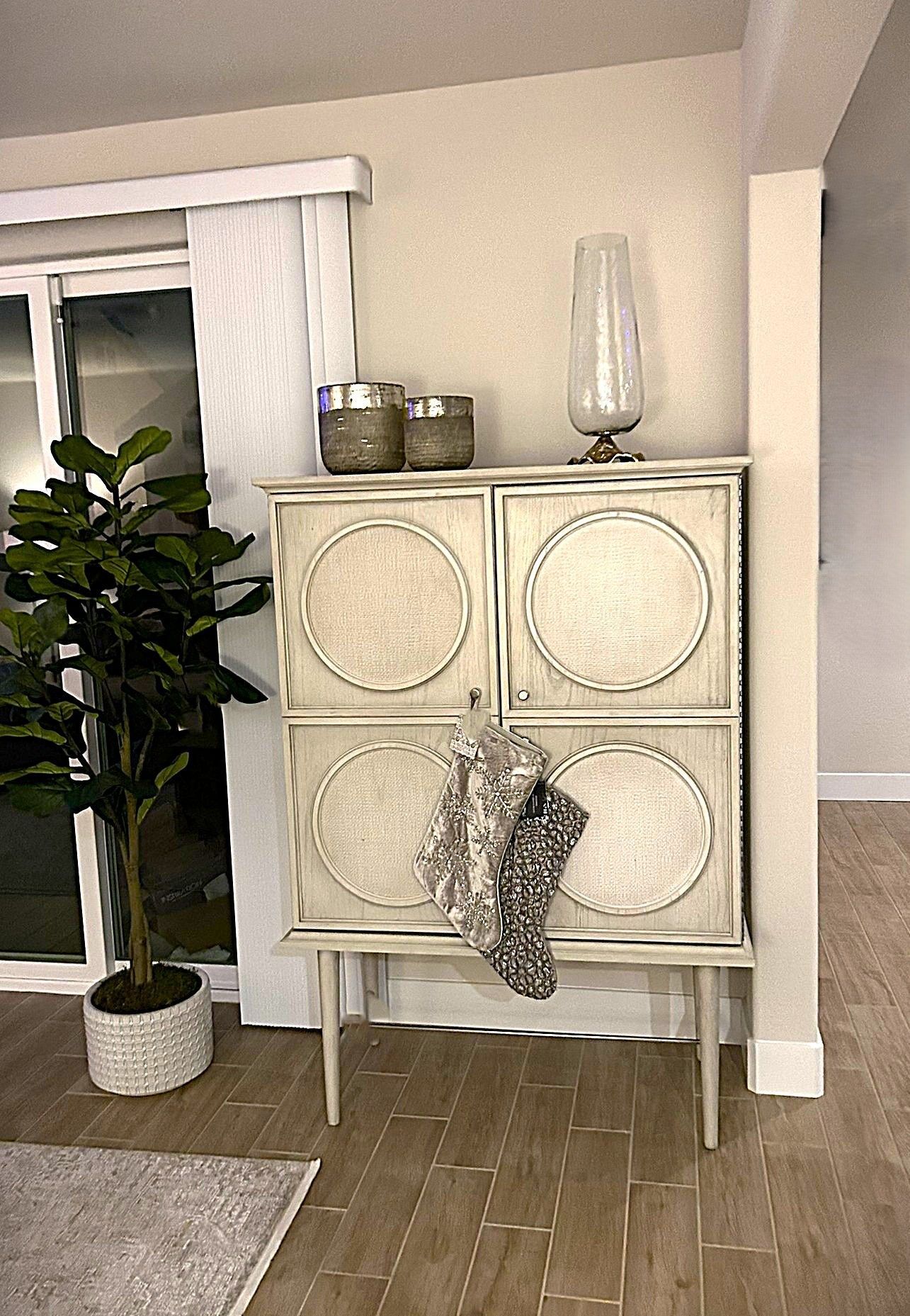 A cream-colored, four-door wooden cabinet with textured circular panels stands near a potted tree and glass vase.