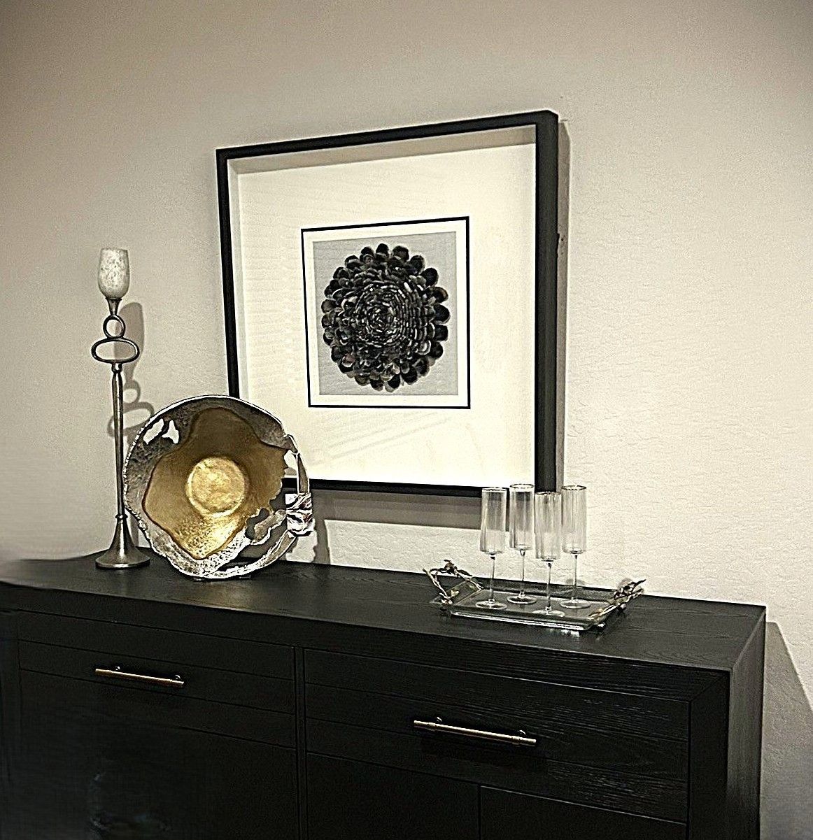 A black dresser topped with a framed floral print, a tall candle holder, a decorative bowl, and glasses on a tray.